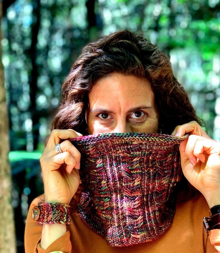 A girl holds a knitted cowl over the lower half of her face to demonstrate the stitch pattern in the knitting pattern