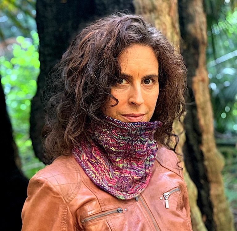 A girl models a cowl made from the Next Chapter Cowl knitting pattern. She has curly hair and stands in front of a burnt out tree in a forest