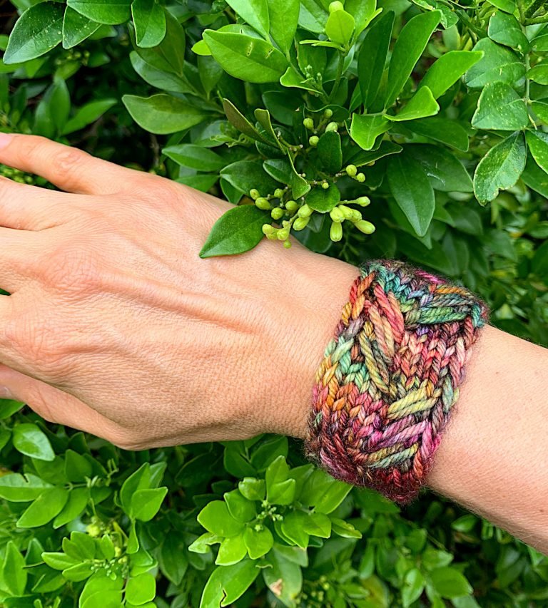 A hand is in front of a green leafy bush with a knitted cuff made from the Next Chapter Cuff around the wrist